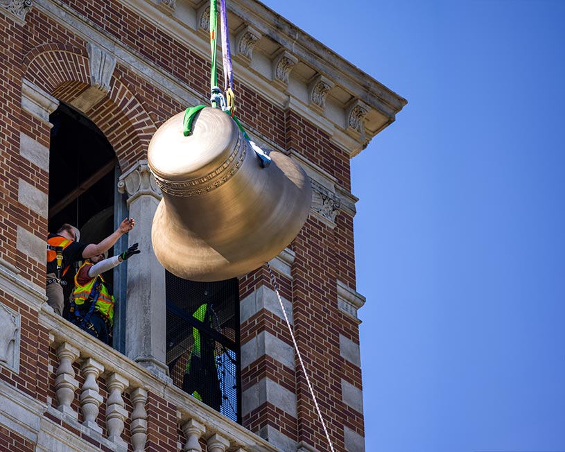 A large bell being lowered into the UNI Campanile tower as workers reach to guide it into the window