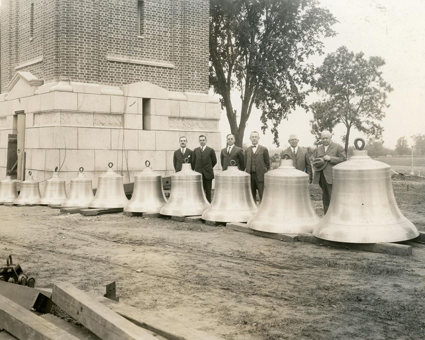 Group of Panther men standing in front of the campanile with the 15-bell chime completed and dedicated and laid out on the ground.