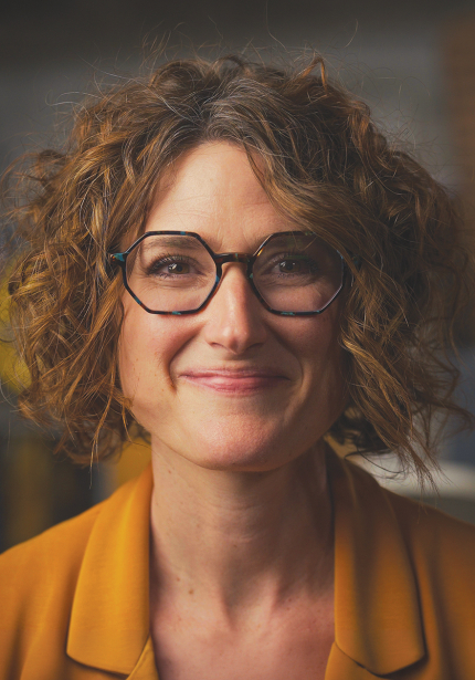 White woman with curly hair and glasses smiling at the camera.